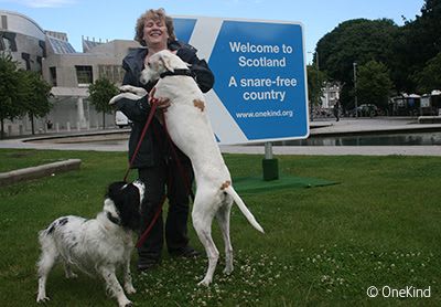 OneKind supporter with dogs beside Snare Free Scotland sign outside Scottish Parliament.